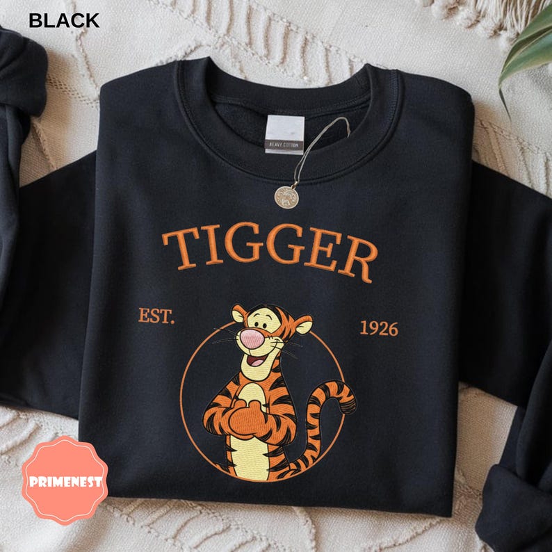 May include: Black sweatshirt featuring the word "Tigger" in orange above an illustration of Tigger from Winnie the Pooh. The sweatshirt also includes "EST. 1926" and a circular graphic of Tigger.