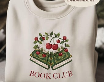 Puede incluir: Sudadera de color crema con un diseño bordado que presenta dos libros verdes, una corona de cerezas y el texto "BOOK CLUB" en rojo. La palabra "EMBROIDERY" está impresa en la parte superior derecha.