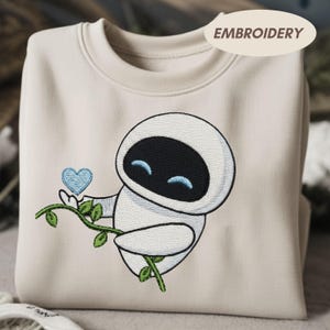 May include: Cream-coloured sweatshirt with an embroidered cartoon robot holding a blue heart and a green vine. The robot has a black face and blue eyes. The word "EMBROIDERY" is printed on the top right of the sweatshirt.