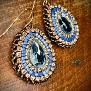 May include: Teardrop earrings featuring a central blue gemstone, surrounded by light blue and gold beads. The earrings have gold-coloured metal hooks, creating an elegant design.