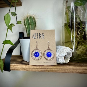May include: A pair of handcrafted earrings with a blue center, surrounded by a white beaded border and a gold-colored metal frame. The earrings are displayed on a brown card with the text "ELK & COVE" and the Instagram handle @elkandcove.