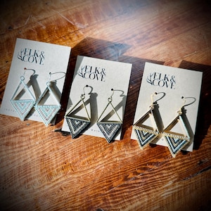 May include: Three pairs of triangle-shaped earrings with geometric beading. The earrings are made of gold-toned metal and feature black, white, and gold beads. The earrings are displayed on three cards with the text "Elk & Cove" printed on them.
