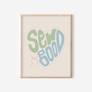 Op de afbeelding: Een ingelijste kunstdruk met de tekst "Sew Good" in een hartvorm. De tekst is groen en blauw, tegen een lichtbeige achtergrond. Een roze hartomtrek en een klein roze strikje met draad zijn ook zichtbaar.