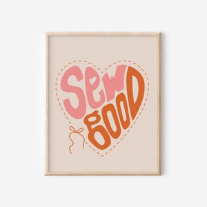 May include: Framed print featuring the phrase "Sew Good" in a heart shape. The words are in pink and orange, set against a light beige backdrop. The heart is outlined with a dashed line, and a small bow and thread are at the bottom.