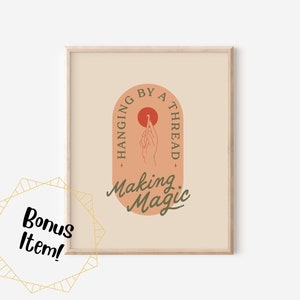 Könnte beinhalten: Ein gerahmter Kunstdruck mit beigem Hintergrund zeigt den Text "Hanging by a Thread" und "Making Magic" in einem stilisierten Design. Das Kunstwerk zeigt eine Hand, die einen Faden hält, und einen roten Kreis. Eine goldene geometrische Form mit den Worten "Bonus Item!" befindet sich in der unteren linken Ecke.