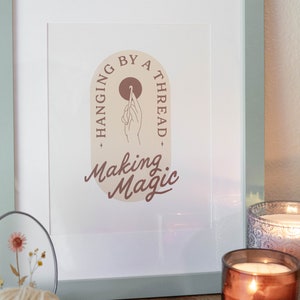 Könnte beinhalten: Gerahmter Kunstdruck mit einem salbeigrünen Rahmen. Der Druck zeigt den Text "Hanging by a Thread, Making Magic" mit einer Handillustration. Ein Wollknäuel, ein dekorativer Teller und eine brennende Kerze befinden sich im Vordergrund.