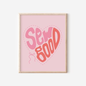 May include: A framed art print featuring a pink background and a heart-shaped design. The heart contains the words "Sew Good" in pink and coral lettering. A small bow and thread detail are at the bottom of the heart.