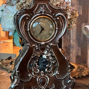May include: A brown and silver mantel clock with an ornate, curved frame and a circular face featuring black Roman numerals. The clock has a dark brown finish with silver highlights along the edges and a small, rounded finial at the top.