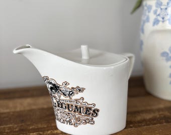 May include: White ceramic creamer with a lid and a spout. The creamer features a decorative black and brown design with the word "LEGUMES" on the side. A vintage-style pitcher is in the background.