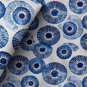 Op de afbeelding: Witte stof met een herhalend patroon van blauwe, gestileerde bloem- of schelpachtige ontwerpen. De blauwe elementen hebben een aquareleffect, met stralende lijnen en een centrale donkerblauwe cirkel. Geschikt voor woondecoratie of kleding.