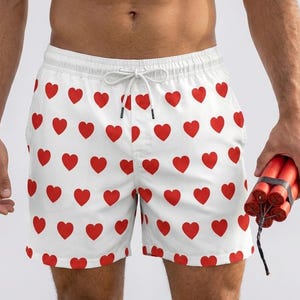 May include: White swim shorts featuring a repeating pattern of small red hearts. The shorts have an elastic waistband with a white drawstring cord. A bundle of red dynamite sticks is held in one hand, positioned to the side of the shorts.