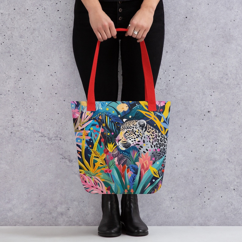 May include: A colorful tote bag with a leopard print design. The bag features a vibrant jungle scene with flowers, leaves, and a leopard's face peeking out from the foliage. The bag has red handles.