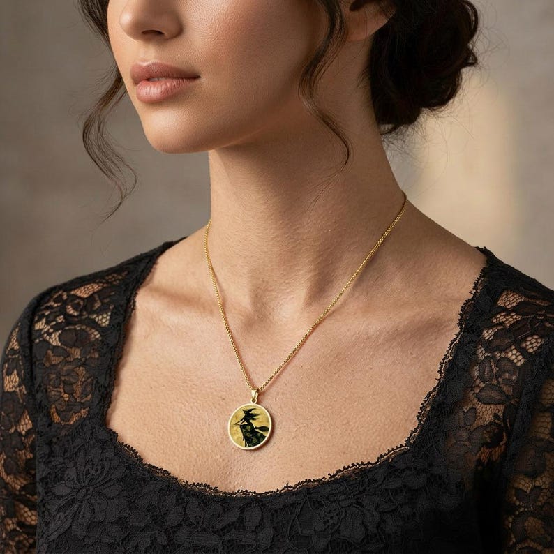 May include: A gold-toned necklace featuring a circular pendant with a black silhouette of a witch flying on a broomstick against a yellow background, suspended from a delicate gold-toned chain resting against a black lace garment.