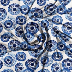 May include: White fabric with a repeating pattern of blue, stylized floral designs. The blue elements have a watercolor effect, with radiating lines from a central dark blue circle. The fabric is slightly wrinkled, suggesting it is draped.