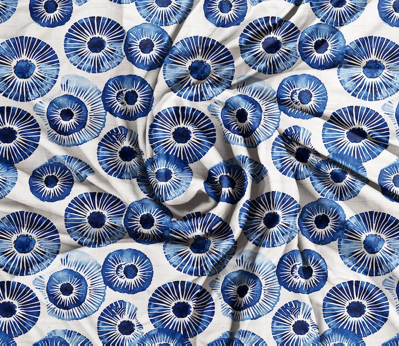 May include: White fabric with a repeating pattern of blue, stylized floral designs. The blue elements have a watercolor effect, with radiating lines from a central dark blue circle. The fabric is slightly wrinkled, suggesting it is draped.