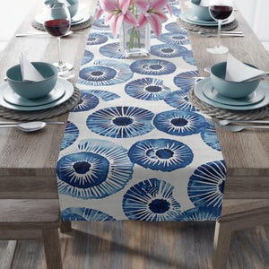May include: A blue and white patterned table runner with a floral design. The runner is placed on a wooden table set for a meal, with plates, bowls, and glasses visible. The runner's design features large, stylized blue floral shapes.