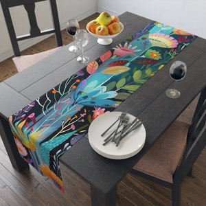 May include: A colorful floral table runner with a dark blue background. The runner features a variety of flowers, leaves, and stems in shades of pink, purple, green, yellow, and orange. The runner is laid out on a dark wooden table with a white plate and silverware.