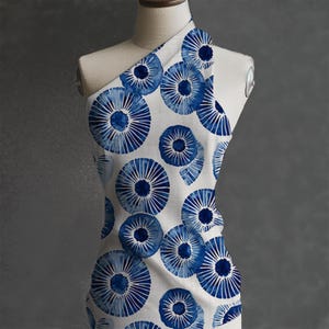 May include: White fabric with a repeating pattern of large, blue, circular designs. Each circle has a dark blue center with radiating lines, resembling stylized flowers or sunbursts. The fabric is draped over a mannequin.