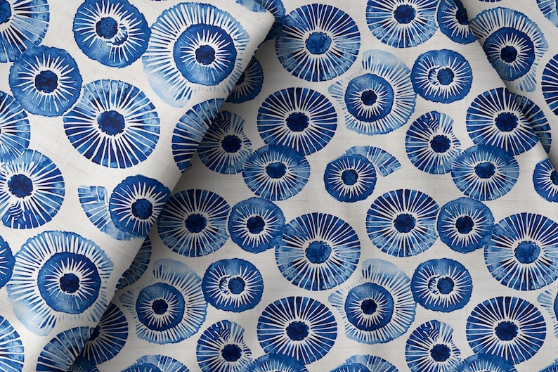 May include: White fabric with a repeating pattern of blue, stylized floral or shell-like designs. The blue elements have a watercolor effect, with radiating lines and a central dark blue circle. The fabric is likely used for home decor or apparel.