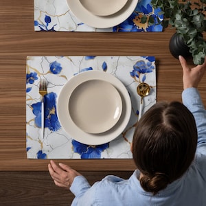 May include: A white dinner plate with a blue floral design placemat. The placemat has a gold pattern and a white background. A gold fork and spoon are on the placemat.