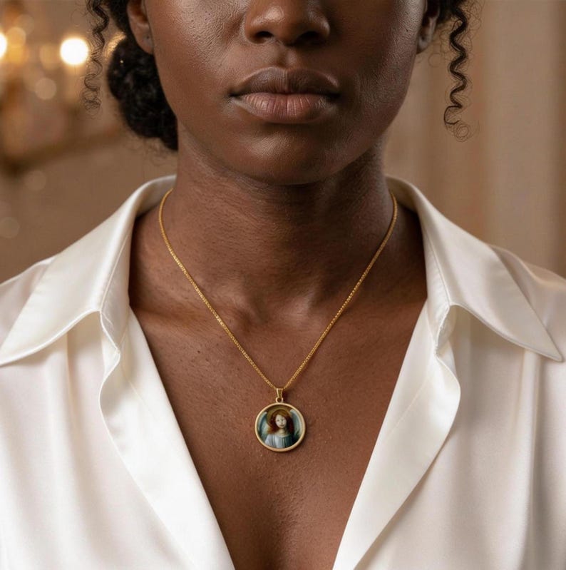 May include: A gold-toned chain necklace featuring a circular pendant with a detailed portrait of a figure with long hair and a halo. The pendant has a thin gold border and hangs from a delicate, textured gold-colored metal chain link necklace.
