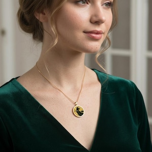 May include: A gold-toned necklace featuring a circular pendant with a black silhouette of a witch flying on a broomstick against a textured gold background, suspended from a delicate gold chain worn against a dark green velvet garment.