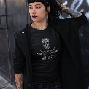 May include: Black t-shirt featuring a decorative skull design and the text "the Vampire Lestat 1984 Tour San Francisco USA". The shirt is paired with a plaid skirt and a black jacket. The overall aesthetic is dark and gothic.