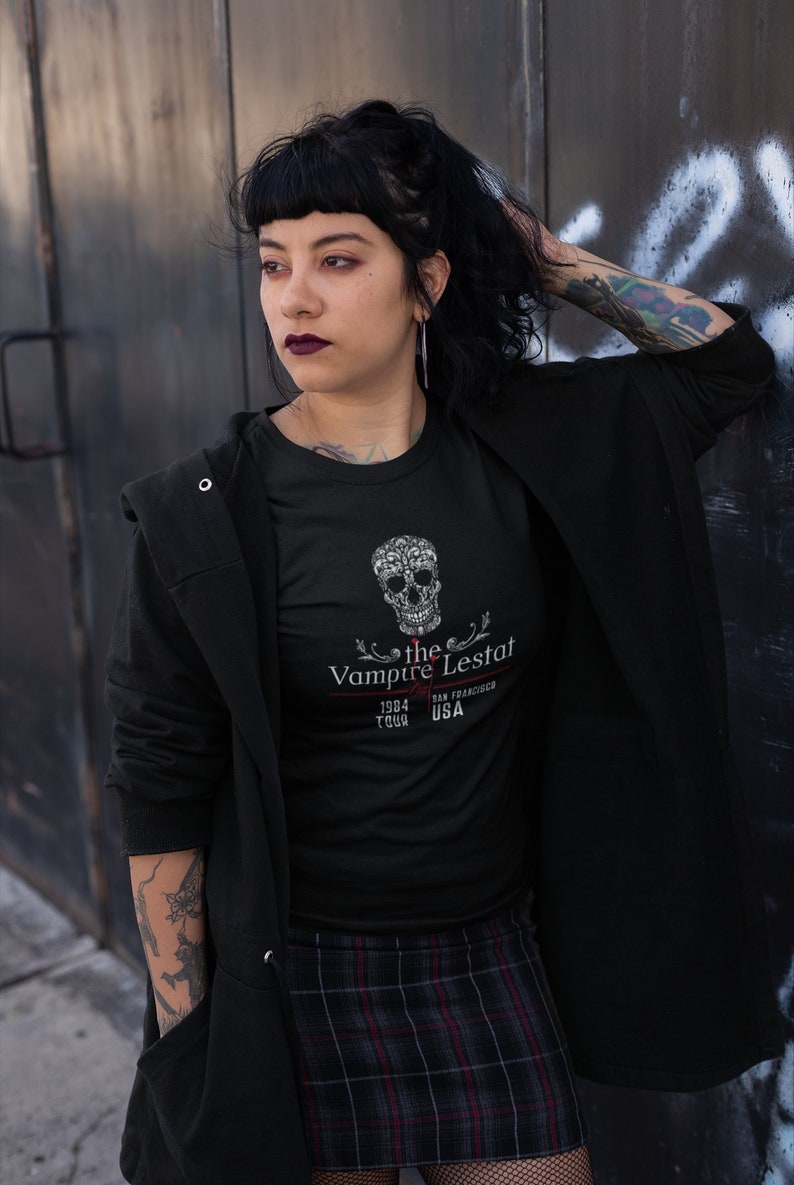 May include: Black t-shirt featuring a decorative skull design and the text "the Vampire Lestat 1984 Tour San Francisco USA". The shirt is paired with a plaid skirt and a black jacket. The overall aesthetic is dark and gothic.