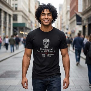 May include: Black t-shirt featuring a decorative skull design with the text "the Vampire Lestat 1984 Tour USA". The shirt is a crew neck style and is made of a soft material. The design is in white.
