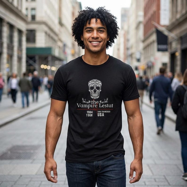 May include: Black t-shirt featuring a decorative skull design with the text "the Vampire Lestat 1984 Tour USA". The shirt is a crew neck style and is made of a soft material. The design is in white.