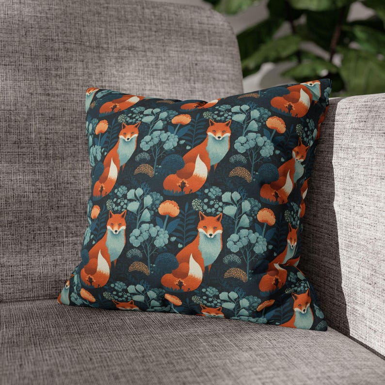 May include: Square throw pillow with a repeating pattern of foxes, flowers, and foliage. The foxes have orange fur and light blue accents, set against a dark teal background. The pillow is placed on a gray sofa.