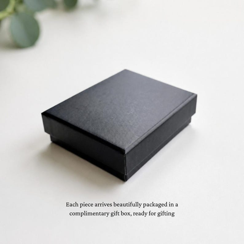May include: A rectangular black gift box with a textured surface finish. The box is closed and features a clean, simple design with sharp edges, presented as a solid container for small items, positioned on a plain, light-colored surface.