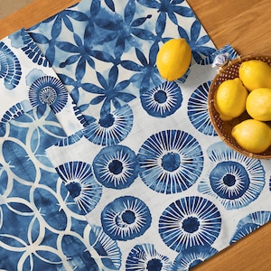 May include: A white tablecloth with a blue and white floral pattern. The tablecloth features a repeating pattern of stylized flowers and circles. The tablecloth is laid out on a wooden table with a bowl of lemons in the background.