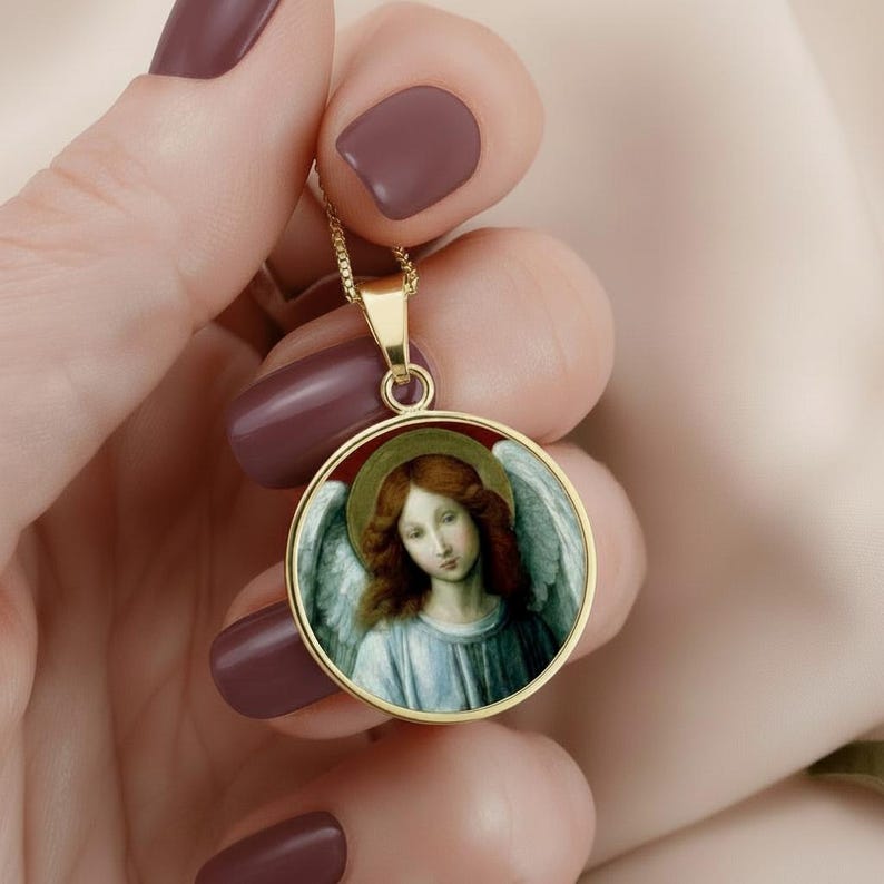 May include: A gold-toned circular pendant necklace featuring a detailed artistic print of an angel with auburn hair, white wings, and a golden halo. The pendant is suspended from a delicate gold-colored chain, showcasing a classic religious motif.