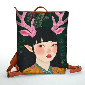 May include: A green canvas backpack featuring a colourful illustration of a woman with pink antlers and a floral pattern. The backpack has brown leather straps and buckles.