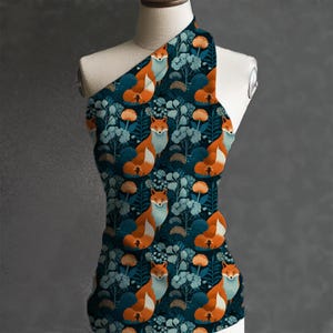 May include: A one-shoulder top featuring a repeating pattern of orange foxes, blue flowers, and orange floral accents on a dark teal background. The fabric is draped over a mannequin, showcasing the design.