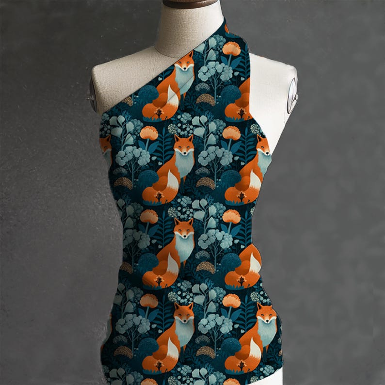 May include: A one-shoulder top featuring a repeating pattern of orange foxes, blue flowers, and orange floral accents on a dark teal background. The fabric is draped over a mannequin, showcasing the design.