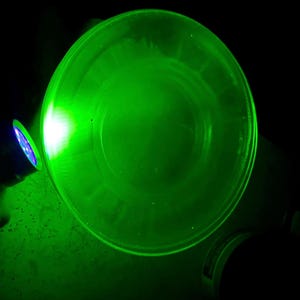 May include: A clear, circular glass object illuminated with bright green light. The light source, a small, round device with blue lights, casts a strong glow across the glass, creating a vibrant, luminous effect against a dark background. The object's edges are clearly defined.