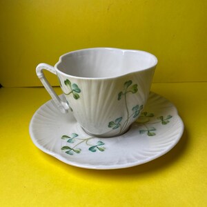 May include: White teacup and saucer set with a shamrock design. The teacup has a handle and is decorated with green shamrocks. The saucer has a scalloped edge and also features shamrocks. The set is on a yellow background.