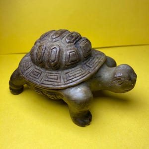 May include: A detailed, carved stone turtle figurine. The turtle has a textured shell with geometric patterns and a detailed head and legs. The figurine is a muted brown color and sits on a yellow background.