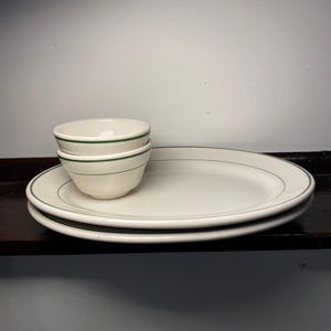 May include: A stack of two small, off-white bowls and two matching oval plates. Each piece has a thin green line around the rim. The dishes are stacked on a dark brown surface.