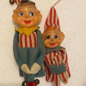 May include: Two vintage elf ornaments. One elf wears a blue jacket with red and white striped shirt and red shoes with bells. The other elf wears a red and white striped outfit and a matching hat. Both have smiling faces and blue eyes.