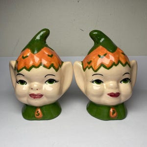 May include: A pair of vintage ceramic salt and pepper shakers shaped like elf faces. The shakers have green caps and collars, orange and cream-colored faces, and are marked with an "S" and a "P".