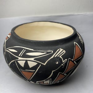 Könnte beinhalten: Eine schwarze Keramikschale mit cremefarbenem Innenraum. Die Außenseite zeigt ein Vogeldesign mit weißen und orangefarbenen geometrischen Formen. Die Schale hat eine runde Form und eine weite Öffnung, die einen traditionellen Keramikstil zeigt.