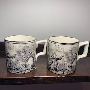 May include: Two white ceramic mugs with a black and white illustration of a historical scene. The mugs have a silver rim and a square handle. The illustration depicts figures in period clothing and furniture.