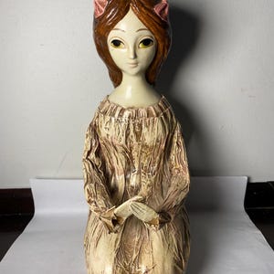 May include: A vintage ceramic figurine of a kneeling woman. The figurine has brown hair with pink bows, large eyes, and is wearing a long, textured dress in shades of beige and brown. The hands are clasped in front.
