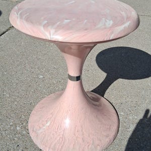 May include: A pink and white swirl-patterned side table with a round top and a pedestal base. The table has a silver band around the middle of the pedestal. The table is made of a glossy material.