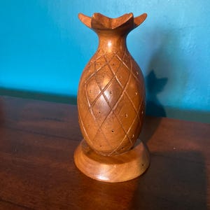 May include: A wooden pineapple-shaped decorative item. The pineapple is brown with a diamond pattern carved into the surface. It has a round base and a crown of leaves at the top. The item is sitting on a dark brown surface.