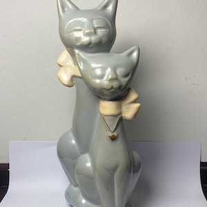 May include: A gray ceramic cat figurine featuring two cats, one behind the other. Each cat has closed eyes and a cream-colored bow. The front cat wears a gold heart pendant. The figurine is a decorative item.