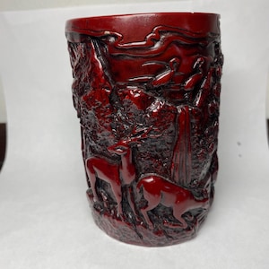 May include: A cylindrical, dark red decorative container with carved relief details. The relief depicts a forest scene with deer and birds. The container has a textured surface and a smooth rim. The overall design is intricate and detailed.